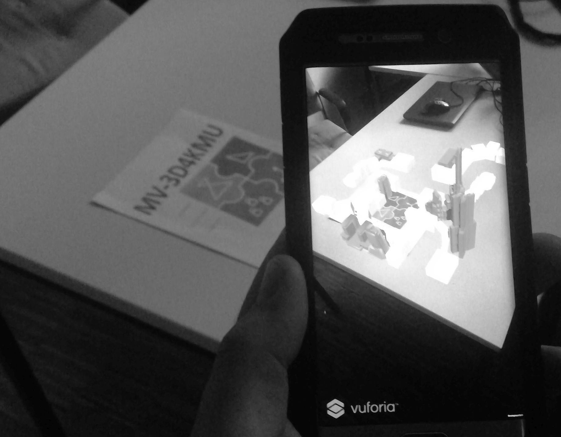 Abbildung 1: Das Bild zeigt ein Handy und einen Tisch auf dem ein A4-Blatt liegt, mit dem Projektlogo 'MV-3D4KMU'. Auf dem Bildschirm befindet sich die Android Anwendung mit dem virtuellen 3D Modell visualisiert im Kamerabild des Handys.